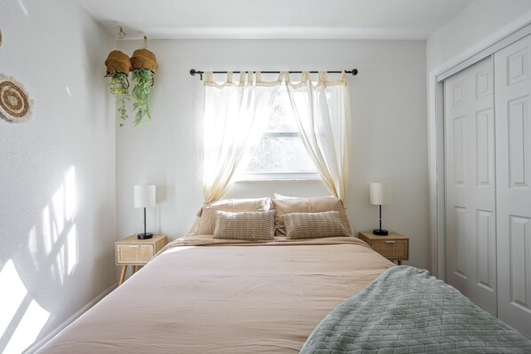Une chambre à coucher bien rangée, avec un grand lit, deux tables de chevet avec des lampes, une fenêtre avec des rideaux légers et une porte.
