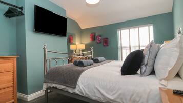 Un dormitorio con paredes color turquesa, una cama con ropa de cama blanca y gris, un aparador de madera, un televisor de pantalla plana y una ventana con persianas.