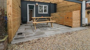 A covered patio area with a wooden bench and a small table, a door leading to a building, and a pebbled ground. - Stillwater Cottage (Beverley)