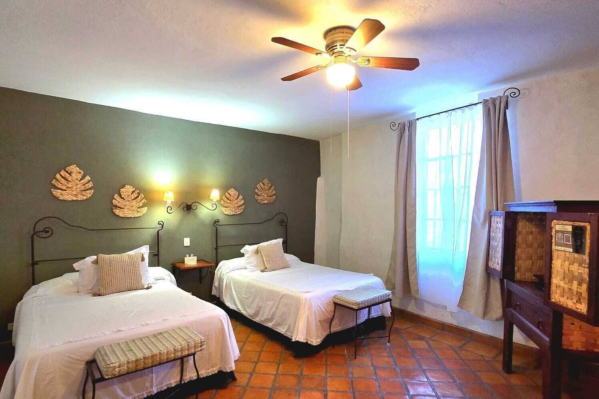 A room with two beds, a ceiling fan, a nightstand with a lamp, and a window with curtains.