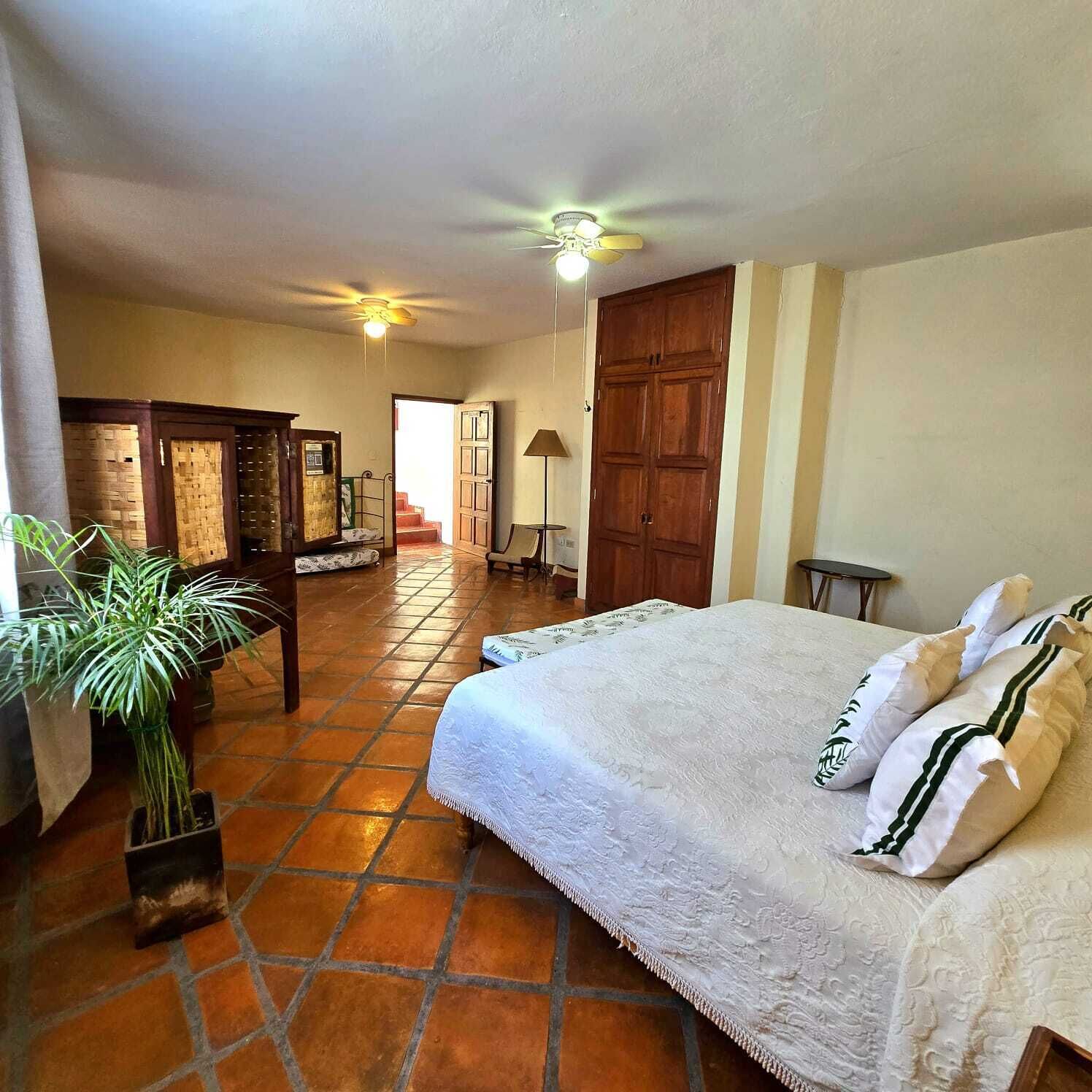 A bedroom with a large bed, a wooden wardrobe, a ceiling fan, and a potted plant.
