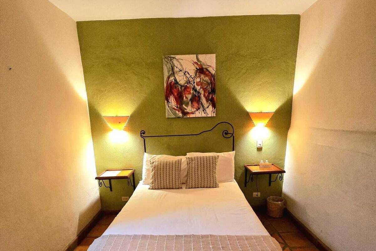 A single-bed room with a green accent wall, two bedside lamps, a painting, and a small bedside table with a lamp.