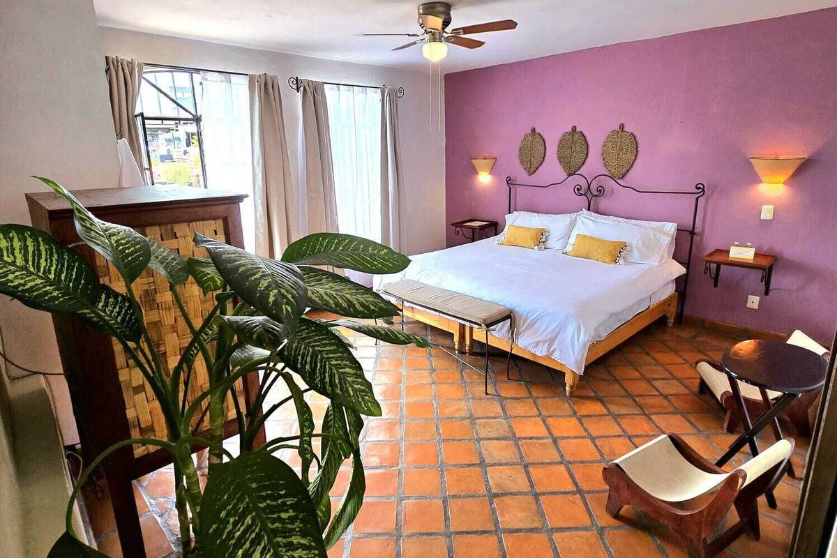 A bedroom with a bed, a ceiling fan, a large plant, a small table, and a chair.