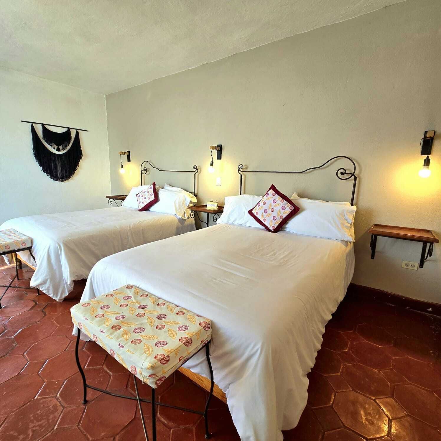 A hotel room with two beds, each with a headboard and bedside tables. There is a bench with a floral cushion in front of one bed.