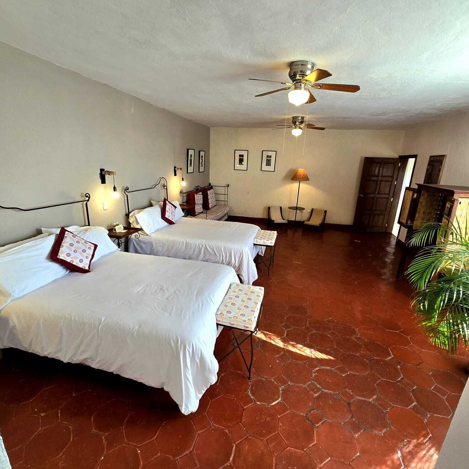 Habitación de hotel con dos camas, ventilador de techo, una mesita, sillas y una puerta.