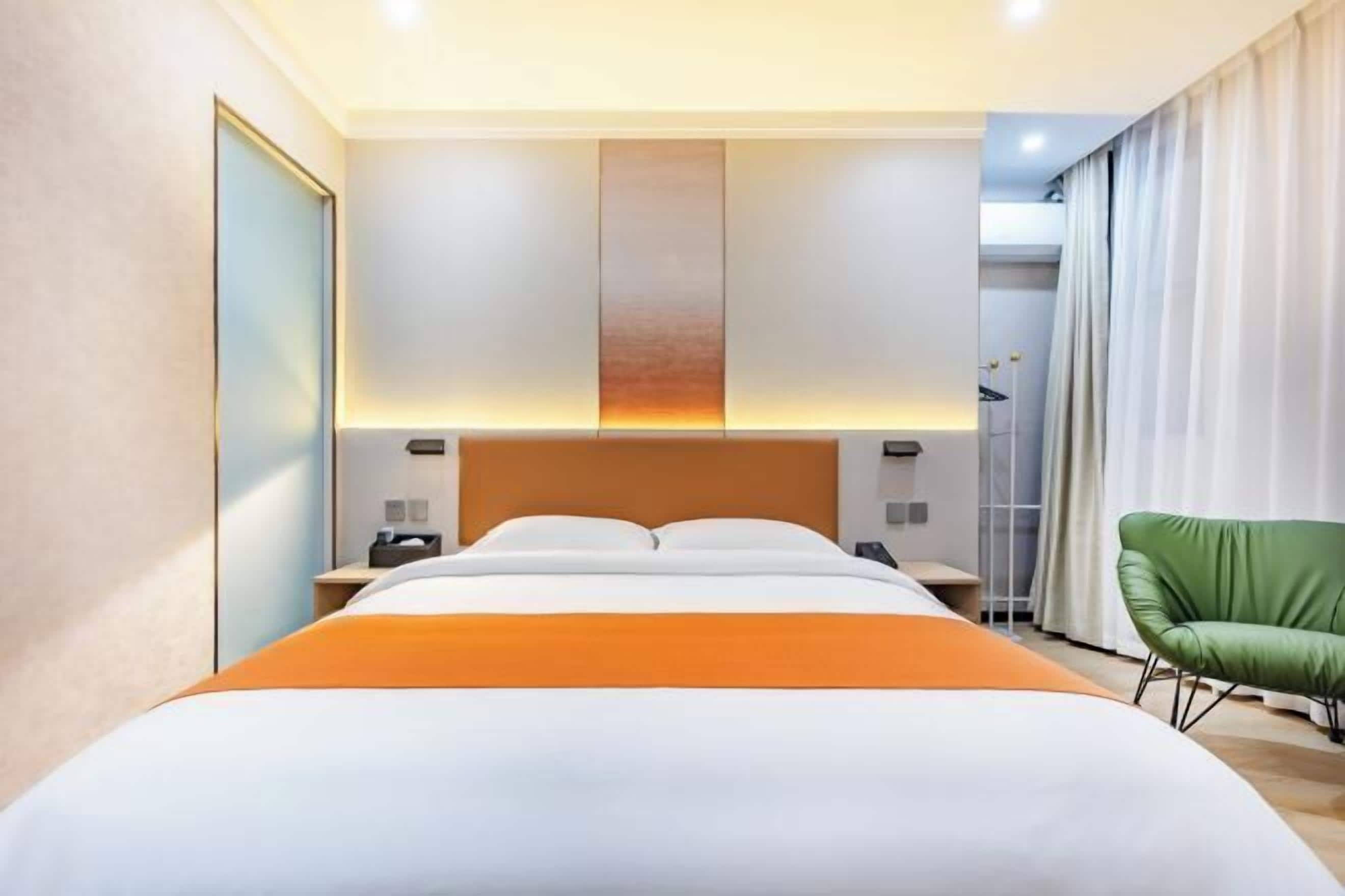 A hotel room with a large bed, a nightstand, a green chair, and a view of the city.