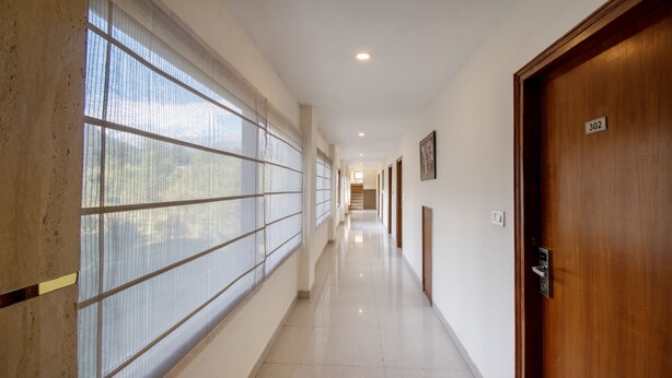 A long, well-lit hallway with a wooden door labeled "302" on the right, a tiled floor, and large windows with vertical blinds on the left. - Treebo Ambika Residency, Near Hidimba Devi Temple (Manali)