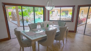 A dining area with a glass table and four chairs, large windows with a view of a pool and garden, and a hanging light fixture.