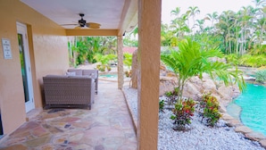 A covered patio with stone flooring, a wicker sofa, and a pool area with tropical plants and a clear blue pool.