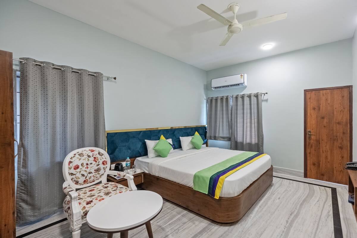 A hotel room with a bed, a chair, a table, a TV, and an air conditioning unit.