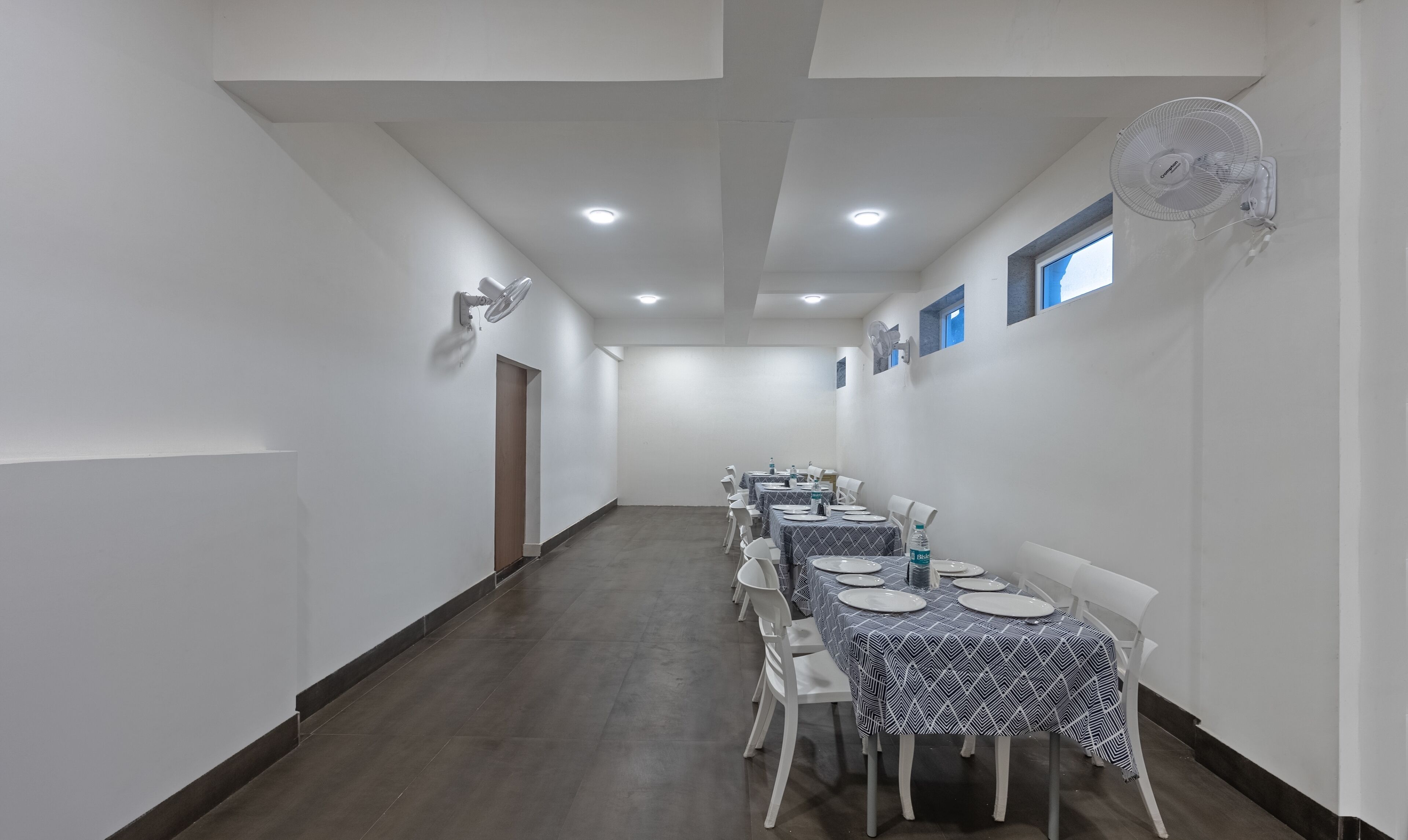A long, narrow room with white walls and a dark floor, lined with tables and chairs. There are ceiling lights and a wall-mounted fan.