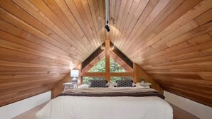 Ein Schlafzimmer mit schrägverdeckter Decke, Holzwänden, einem Bett mit weißer Bettwäsche, zwei Nachttischen mit Lampen und einem Fenster mit Blick auf Grünflächen.