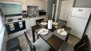 A compact kitchen with white cabinets, a stove, microwave, and a refrigerator. There is a dining table set with plates and napkins.