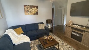 A living room with a dark blue sofa, a coffee table, a patterned rug, a ceiling fan, and a kitchen area with a stove and cabinets.