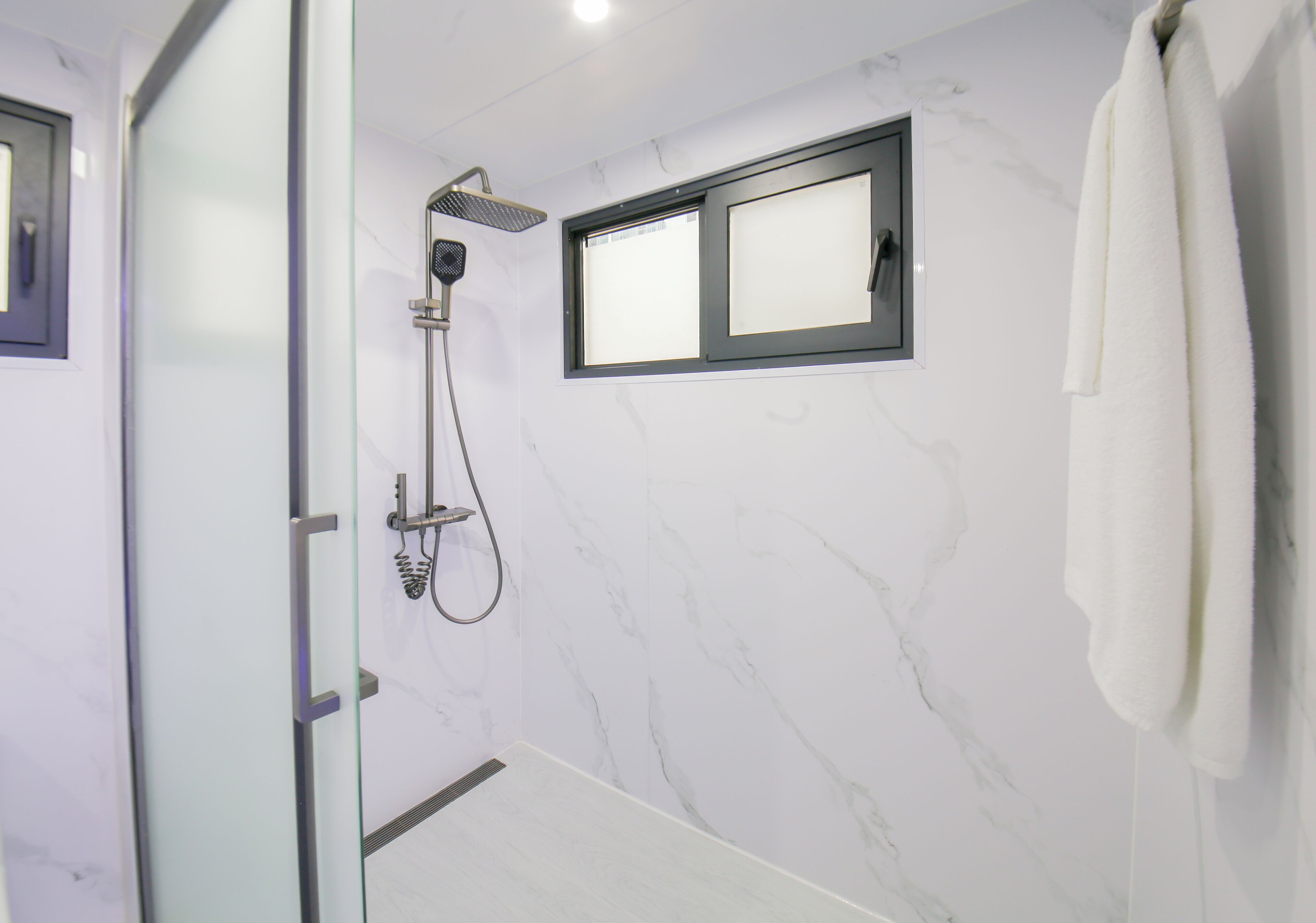 A modern bathroom with a glass shower enclosure, a window with frosted glass, and a white towel hanging on the wall.