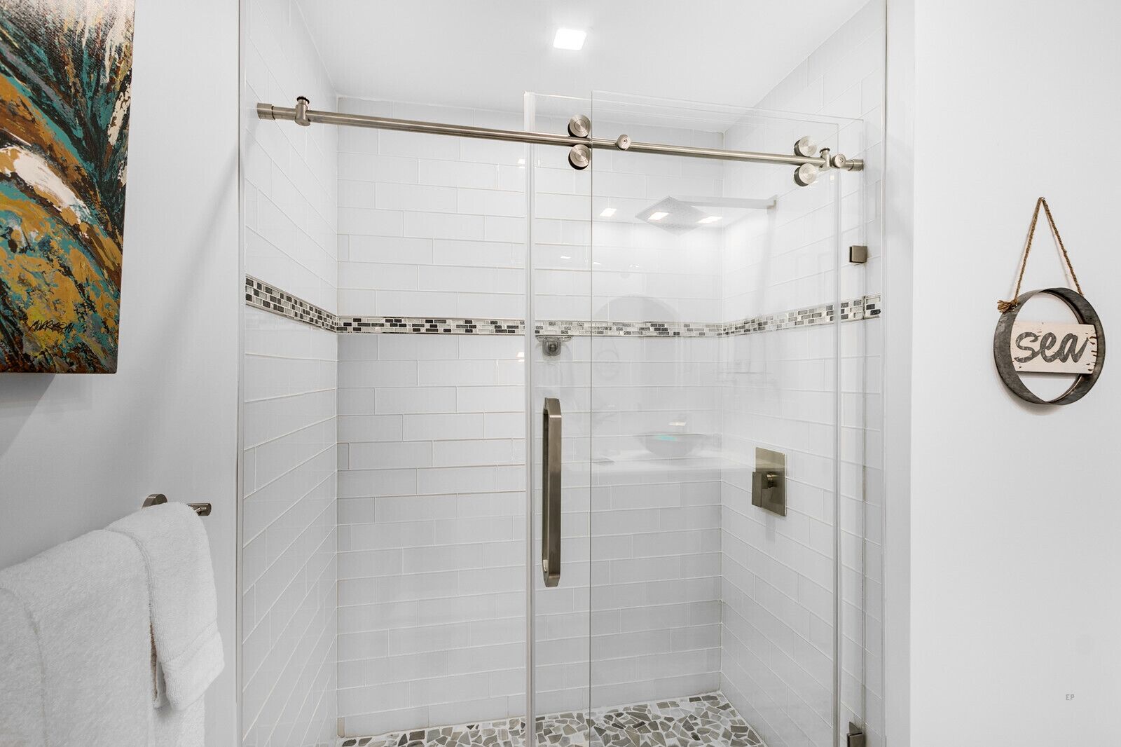 Ducha con cerramiento de vidrio, azulejos blancos y barra de apoyo metálica.
