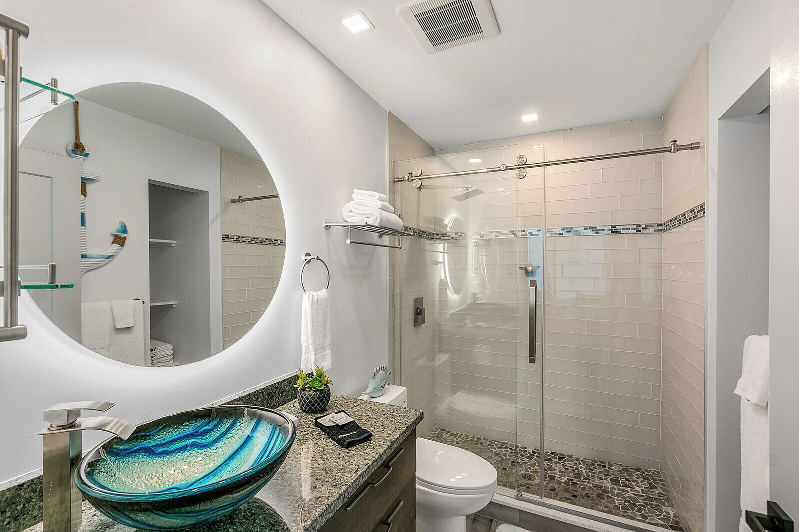 Un baño moderno con ducha de vidrio, un espejo redondo grande y un lavamanos de vidrio azul único.