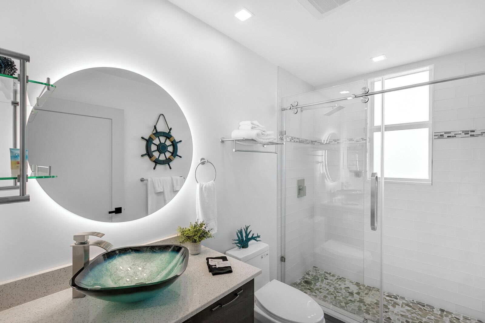 A modern bathroom with a round mirror, a glass sink, a toilet, and a glass-enclosed shower.