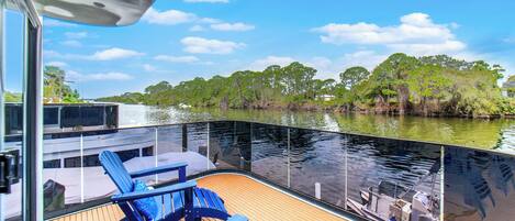 A boat deck with blue chairs and a table, overlooking a calm river with trees and a clear sky.