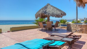 Eine Terrassenanlage am Strand mit Liegestühlen, einem Tisch und einem Reetdachschirm.