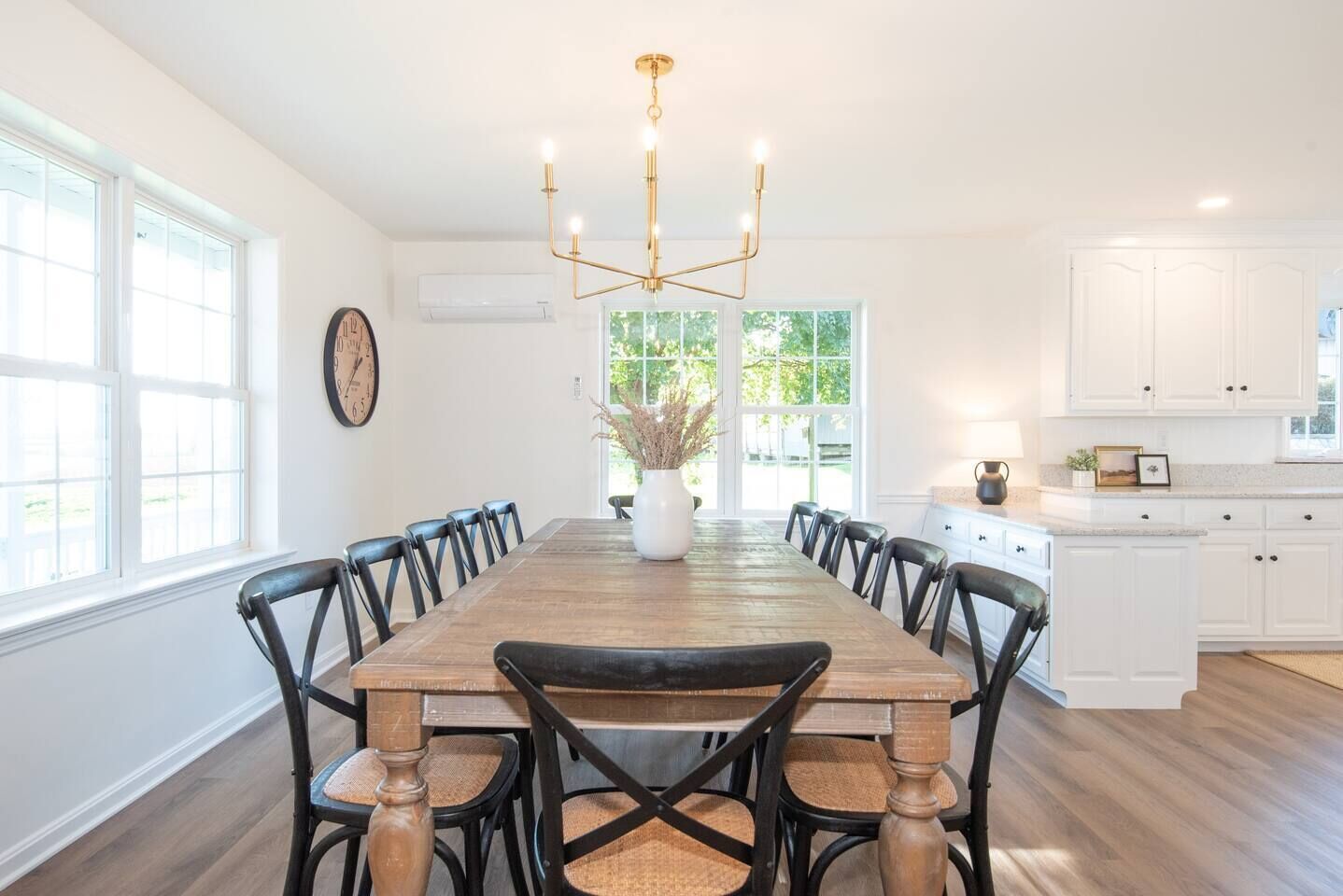 A dining room with a wooden table and black chairs, a chandelier, and a view of the outdoors through large windows.
