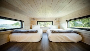 A cabin with two beds, wooden walls, and large windows overlooking a natural landscape. - Dune Cabin - The Dune Cabin is well-appointed and uniquely designed and comfortably sleeps 6 guests. Private bedroom with queen bed, open loft with twin beds, and dinette that converts to a full size bed (Coos Bay)