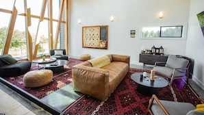 A modern living room with a brown leather sofa, a round coffee table, and a gray armchair. There is a large window with wooden beams, a wall-mounted chessboard, and a small side table with a lamp. - Dune Cabin - The Dune Cabin is well-appointed and uniquely designed and comfortably sleeps 6 guests. Private bedroom with queen bed, open loft with twin beds, and dinette that converts to a full size bed (Coos Bay)