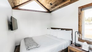 Ein Schlafzimmer mit Holzboden, einem Bett mit weißer Bettwäsche, einem Nachttisch mit Lampe und einem Fenster mit Blick ins Freie.
