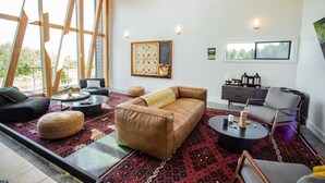 Ein modernes Wohnzimmer mit einem braunen Ledersofa, einem runden Couchtisch und einem grauen Sessel. Es gibt ein großes Fenster mit Holzbalken, ein an der Wand befestigtes Schachbrett und einen kleinen Beistelltisch mit einer Lampe.