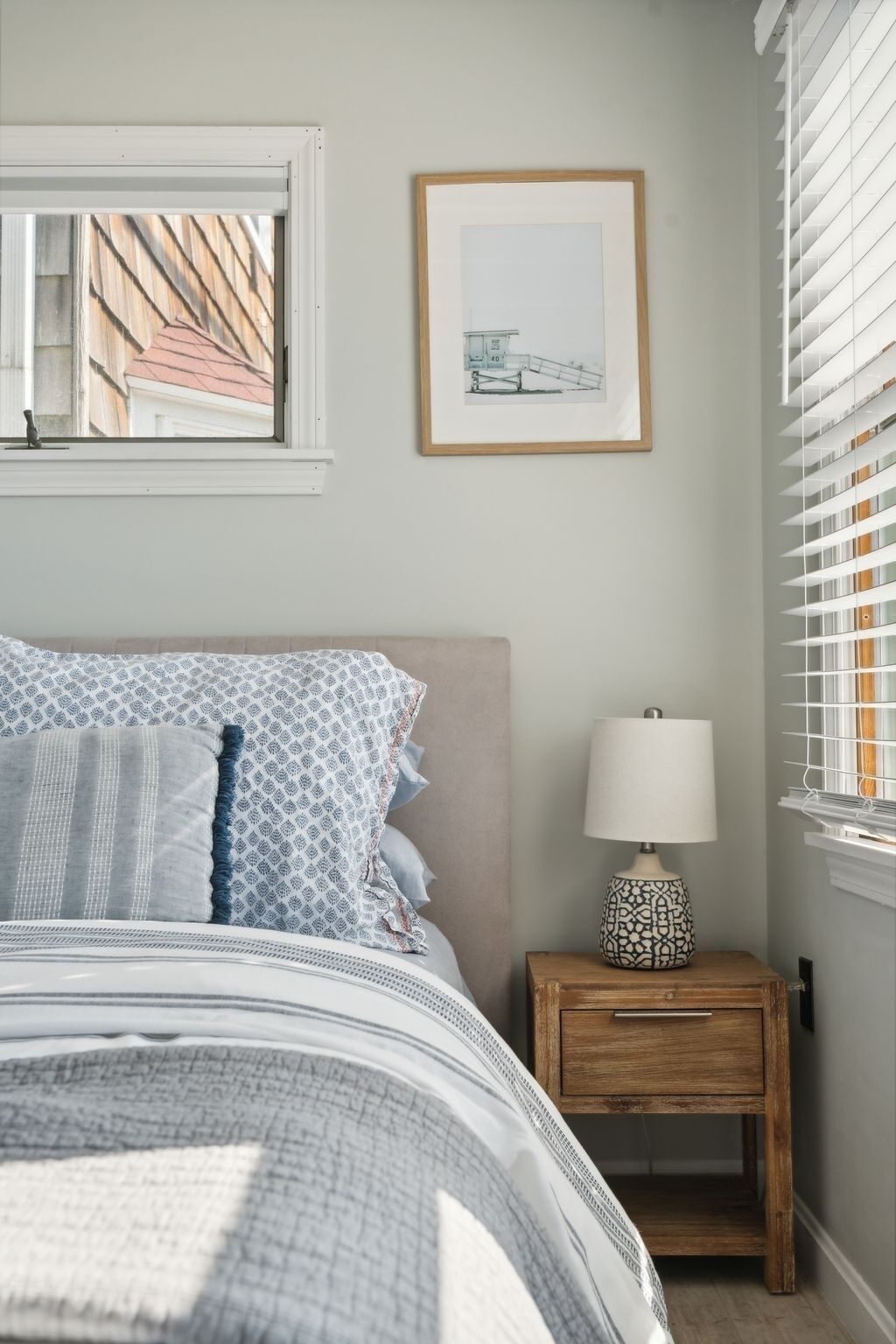 Un dormitorio con cama, una mesita de noche con lámpara, un cuadro enmarcado en la pared y una ventana con persianas.