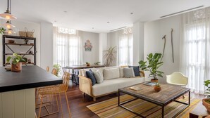 A modern living room with a sofa, coffee table, and chairs. There is a dining area with a table and chairs. The room has large windows with sheer curtains.