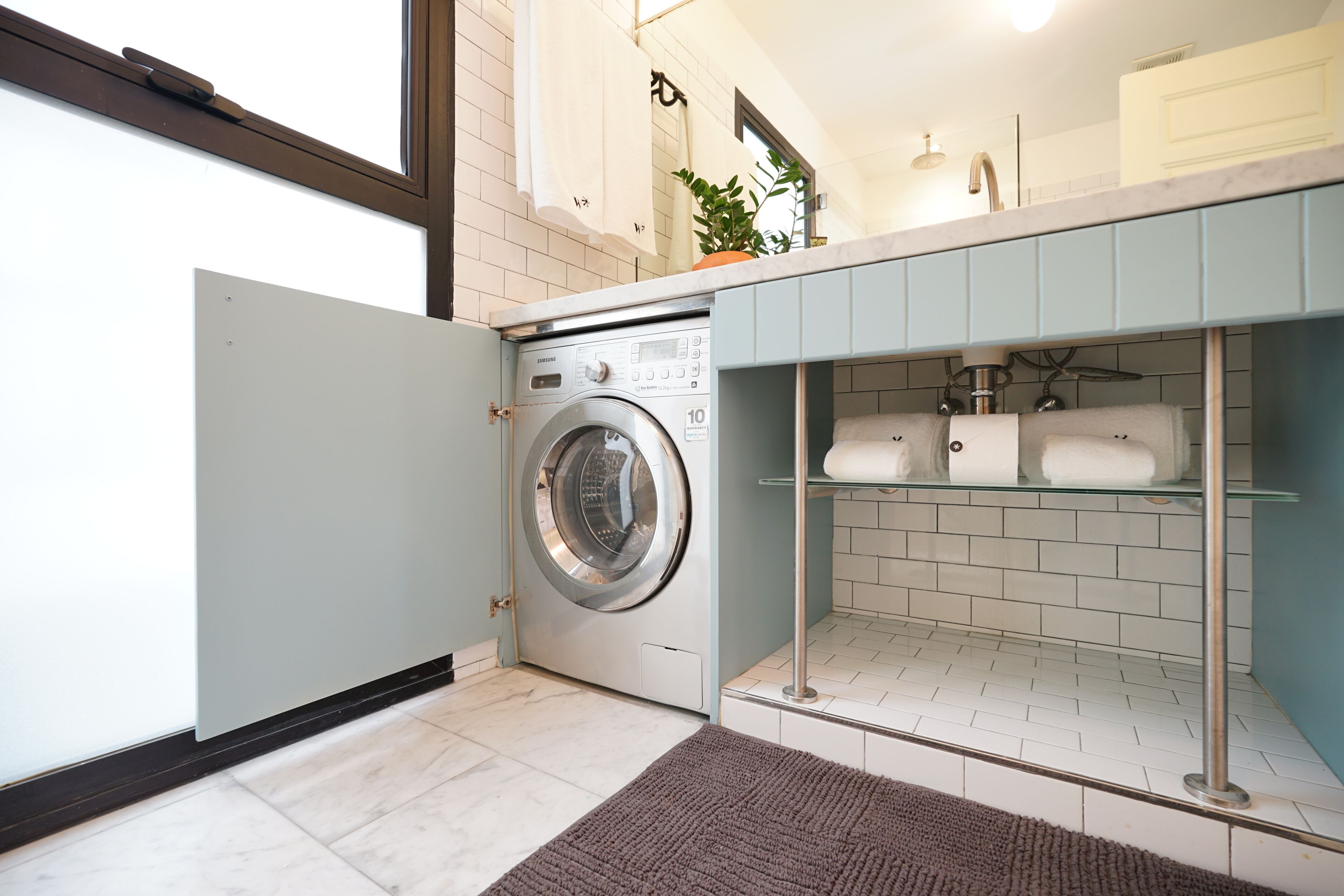 A compact laundry area with a front-loading washing machine, a glass shelf with towels, and a small potted plant.
