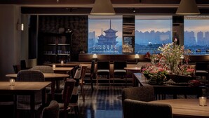 Un restaurante con vistas a un horizonte urbano, mobiliario de madera y un jarrón grande con flores.