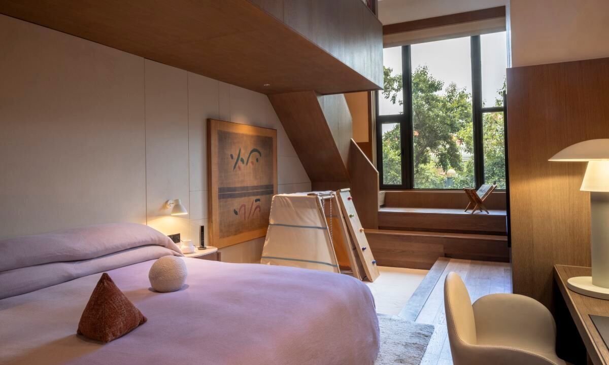 Una habitación de hotel moderna con una cama grande, un escritorio y una silla. Se ve una ventana con vistas a árboles y una escalera.
