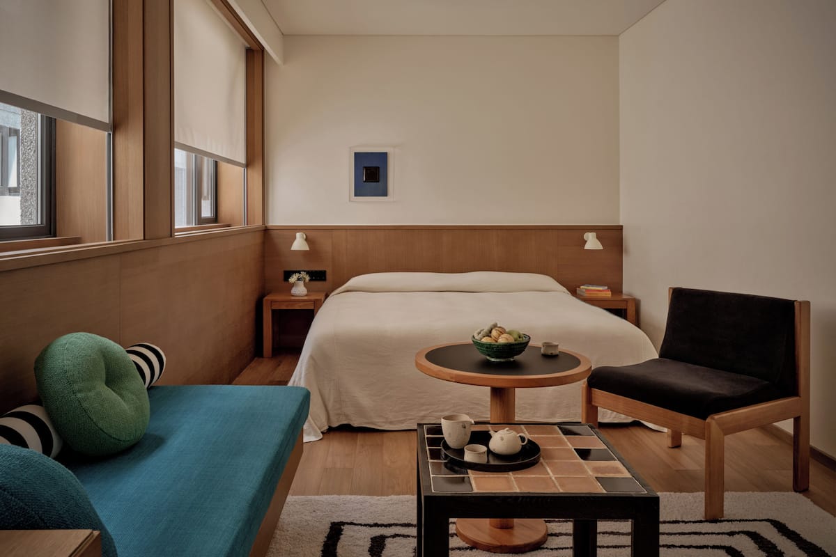 Hotellihuone, jossa on sänky, pieni pyöreä pöytä, jossa on kulho täynnä hedelmiä, tuoli, sohva ja yöpöytä, jossa on lamppu.