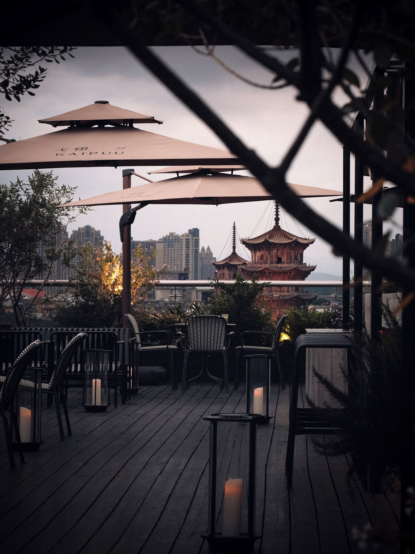 Een houten terras met buitenmeubilair, een pagode-achtige constructie op de achtergrond en een stadsgezicht.
