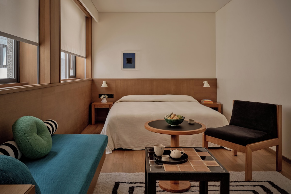 Una habitación de hotel con una cama, una mesita redonda con un frutero, una silla, un sofá y una mesita de noche con una lámpara.