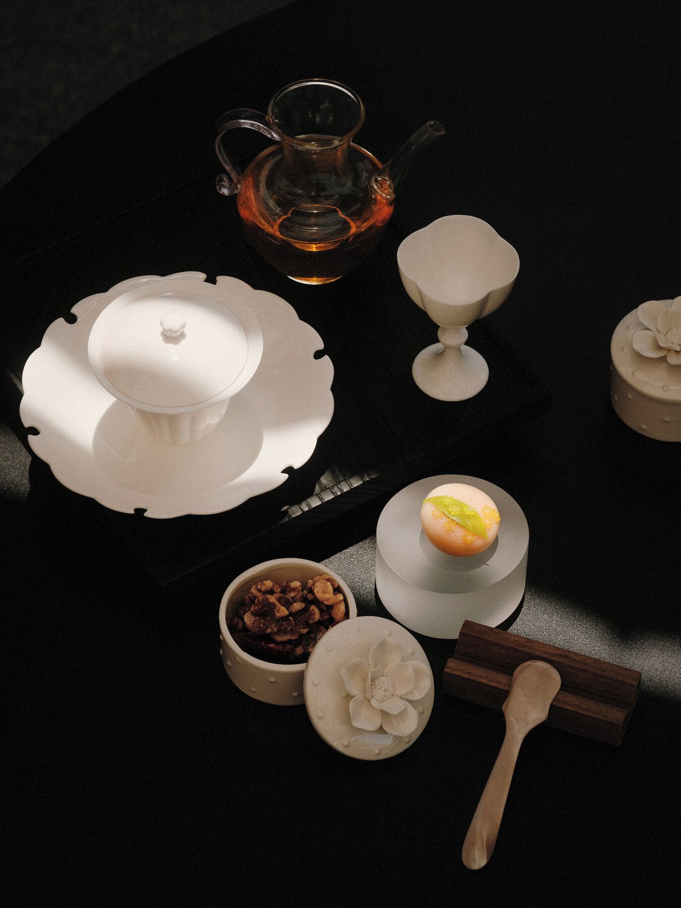 Uma mesa com um bule, uma xícara, uma tigela de nozes, uma colher e um pequeno prato com desenho floral.