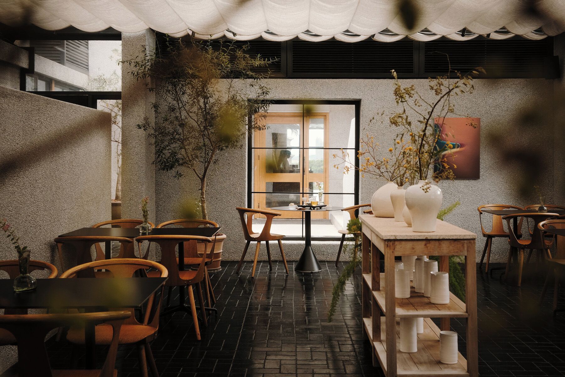 An outdoor seating area with wooden chairs and tables, potted plants, and a view through a glass door.