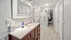 Um corredor estreito com uma porta branca no final, um espelho sobre uma toucador, e um lustre acima do espelho.