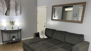 Un canapé gris doté d’un compartiment de rangement, une petite table d’appoint avec deux lampes et une plante, et un grand miroir accroché au mur.