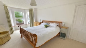 A bedroom with a wooden bed, a nightstand, a chair, and a window with curtains. - Large house (Norfolk) | Sleeps12 | 2min walk to beach | Dogs poss (2) | Playroom (SHERINGHAM)