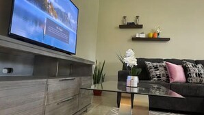 Een moderne woonkamer met een flatscreen televisie bevestigd aan een houten kast, een glazen salontafel, een zwarte bank met decoratieve kussens, en een kleine plant.