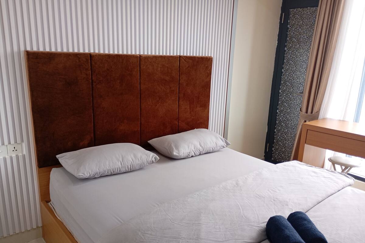 Ein Hotelzimmer mit einem großen Bett, zwei Kissen, einem Holzkopfteil, einem Nachttisch und einem Fenster mit Vorhängen.