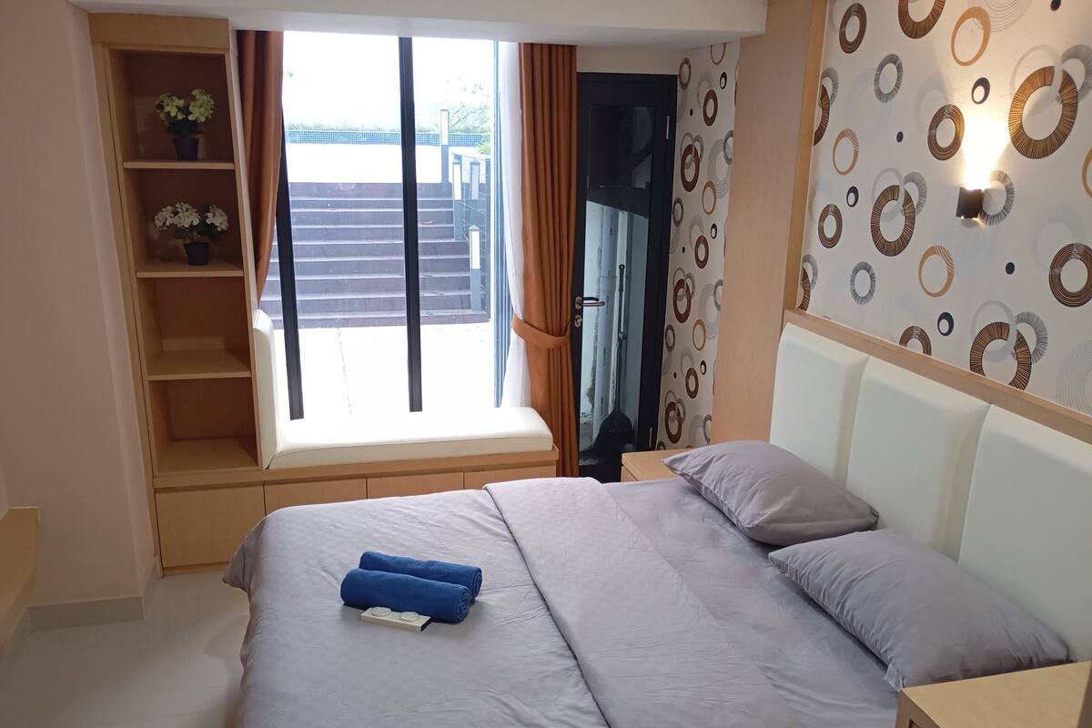 Ein Schlafzimmer mit Bett, Nachttisch, einem Fenster mit Vorhängen, einem Regal mit Pflanzen und einer Wand mit Muster.