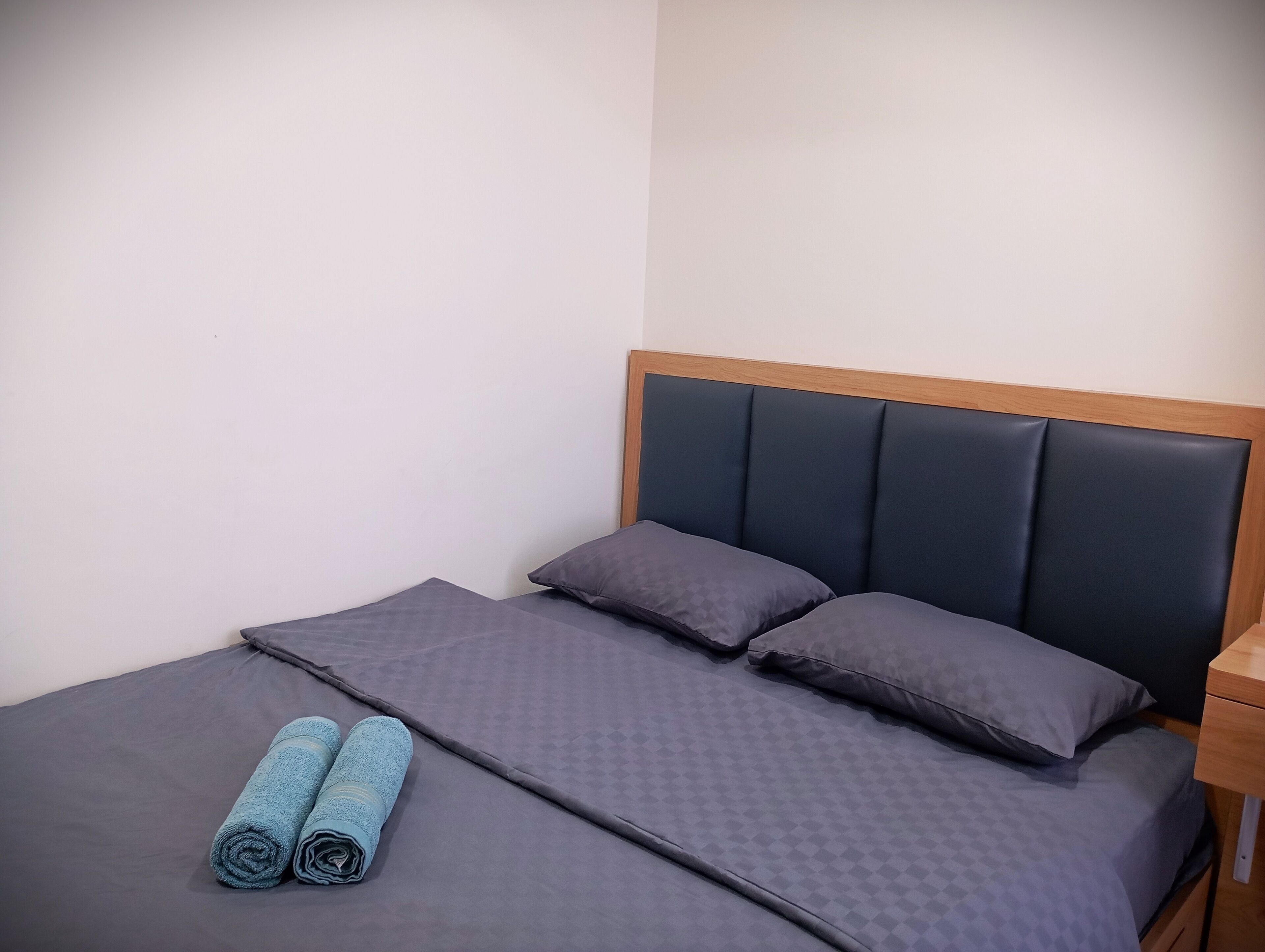 A neatly made bed with a dark headboard and two pillows, a wooden nightstand to the right, and two rolled towels on the bed.