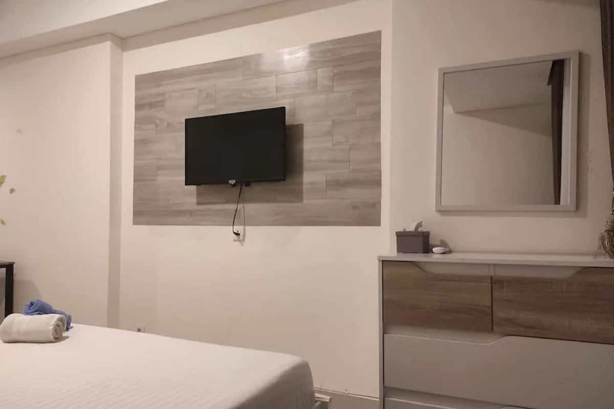 Ein Hotelzimmer mit einem Bett, einem flachbildfernseher an der Wand, einem Schminktisch mit Spiegel und einem kleinen Tisch mit Lampe.
