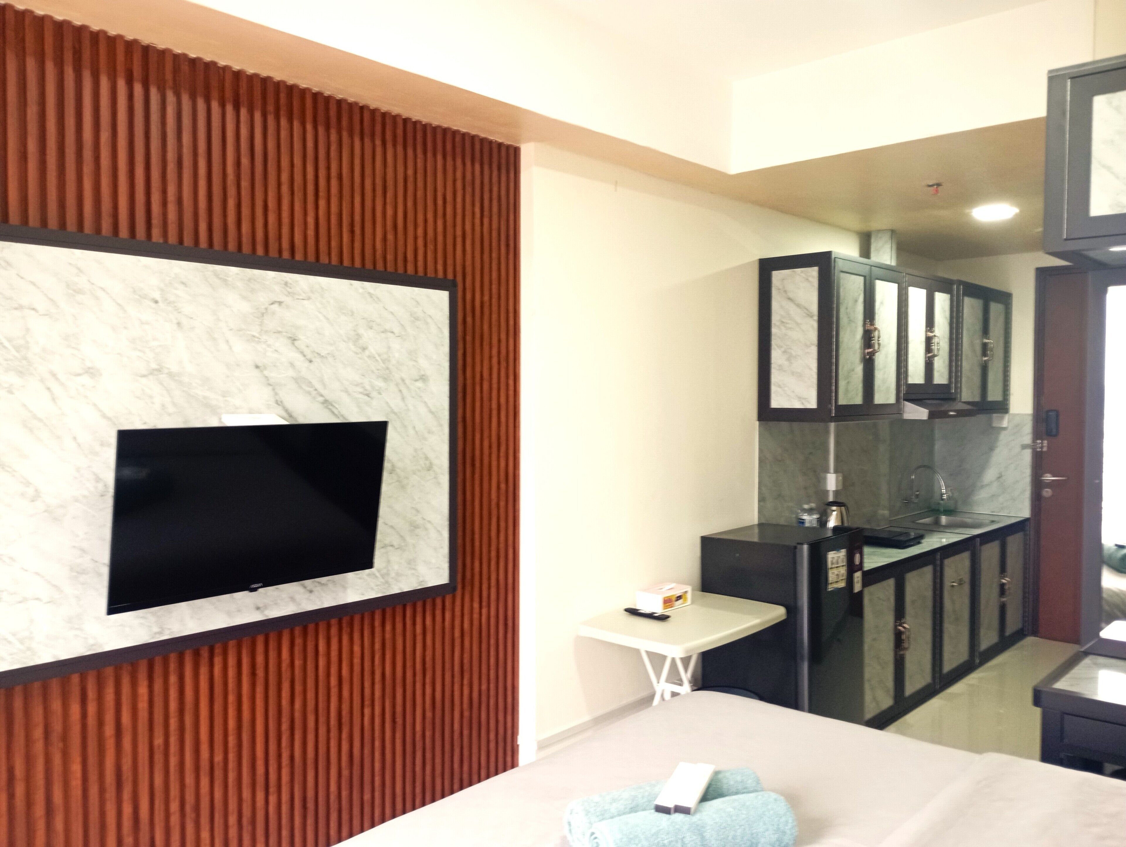 A modern hotel room with a flat-screen TV mounted on a marble wall, a small bedside table, a bed with a blue towel, and a kitchenette with dark cabinets and a marble countertop.