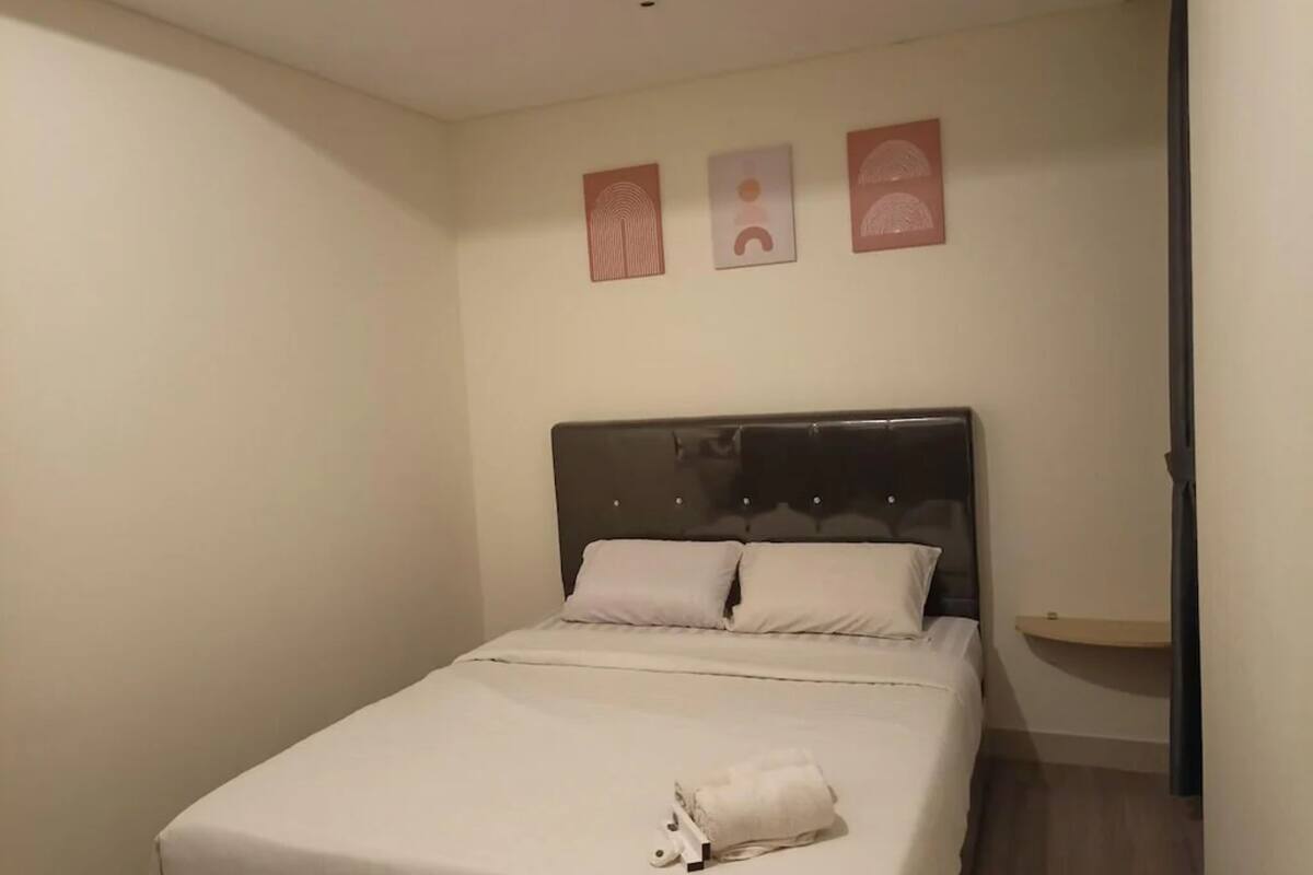 Ein ordentlich bezogenes Bett mit dunklem Kopfteil, einem kleinen Nachttisch und drei an der Wand hängenden Kunstwerken.