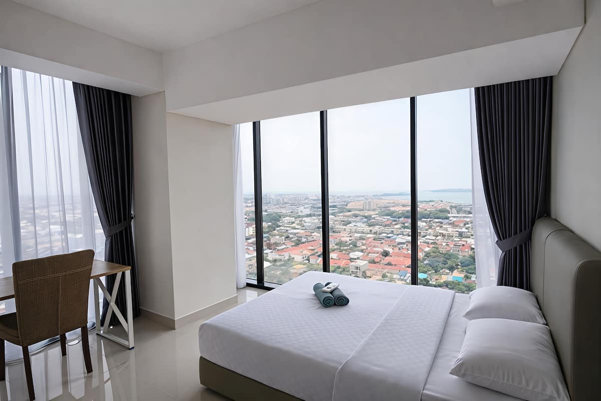 Ein modernes Hotelzimmer mit einem großen Bett, einem Schreibtisch und Blick auf eine Stadtlandschaft durch große Fenster.